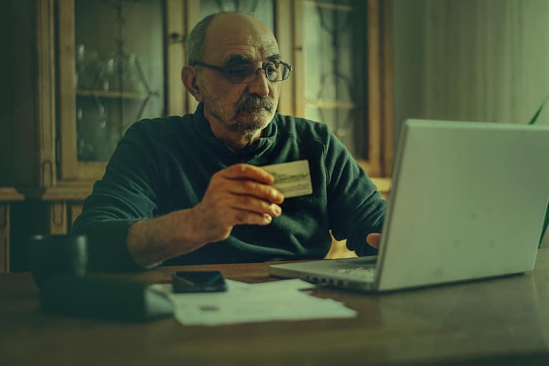 Senior man using a laptop and a credit card for online paying while sitting in his office at home.